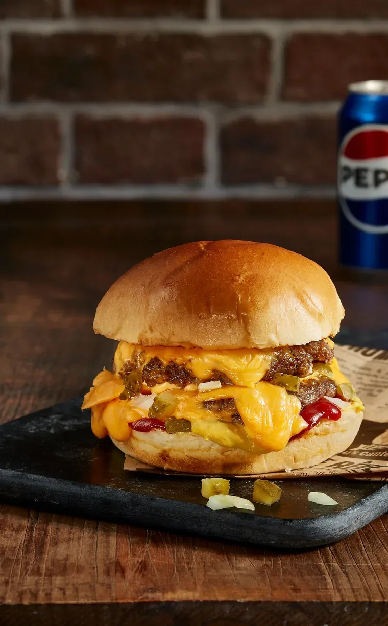 Burger double au cheddar fondu, pickles et oignons, devant une canette